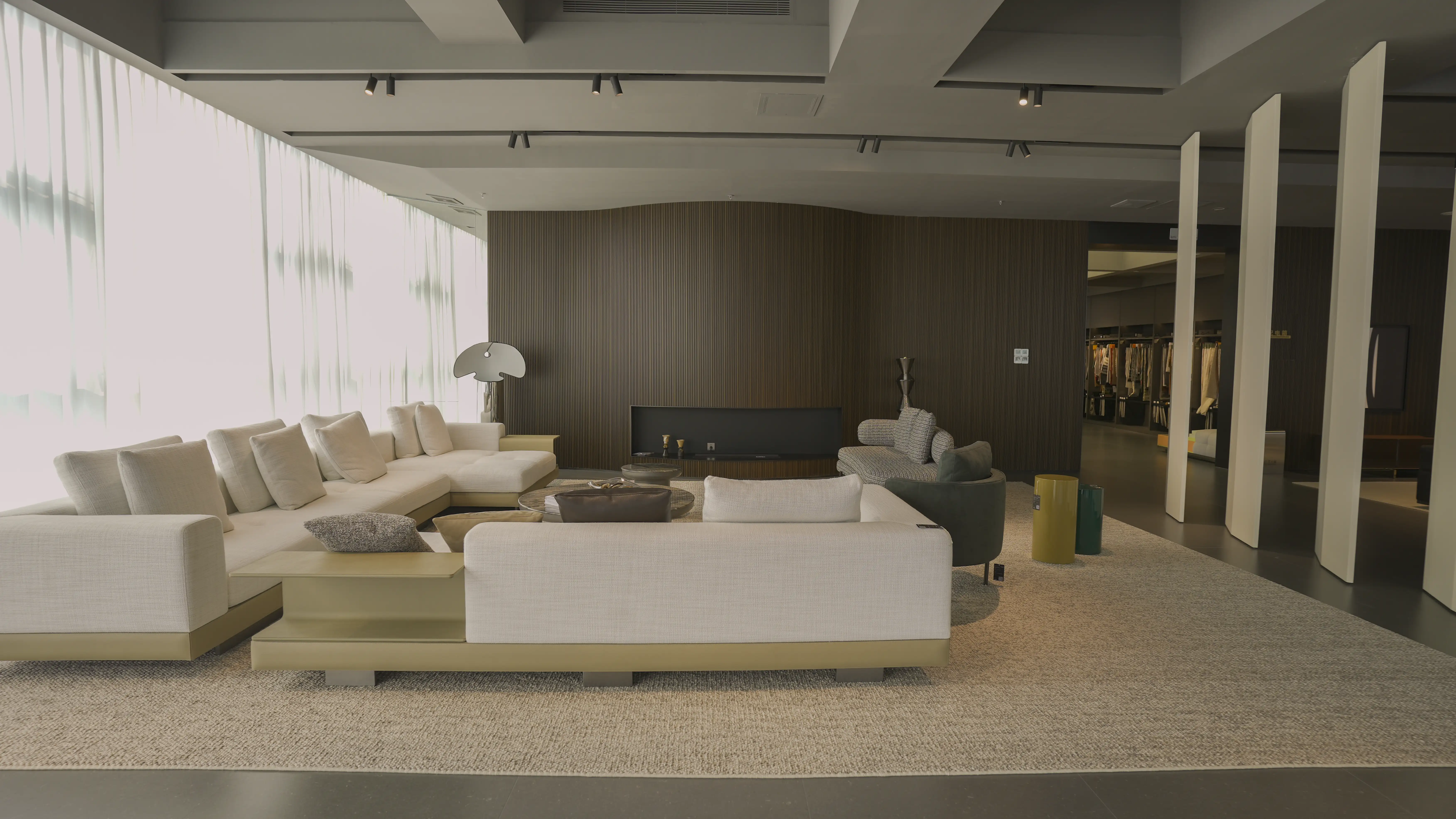 Showroom seating area with cream sofas, gray armchairs, a beige rug, a dark slatted wall and sheer window curtains
