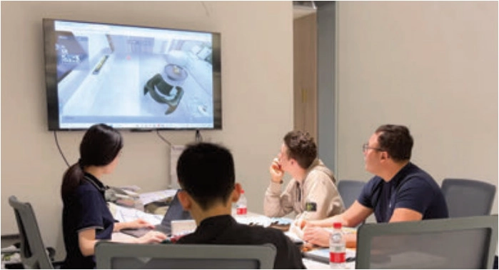 Four people seated at a meeting table watching an interior scene on a wall-mounted screen