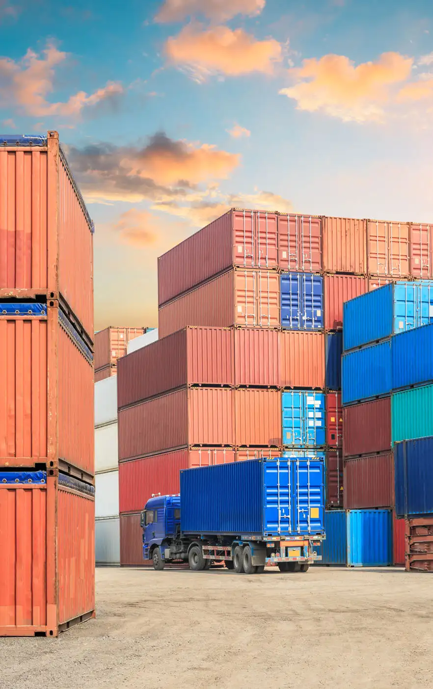 Truck carrying a blue container between stacks of shipping containers beneath a sunset sky