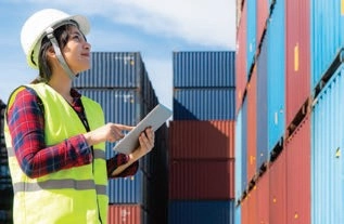 Worker in a hard hat and reflective vest using a tablet beside stacked shipping containers